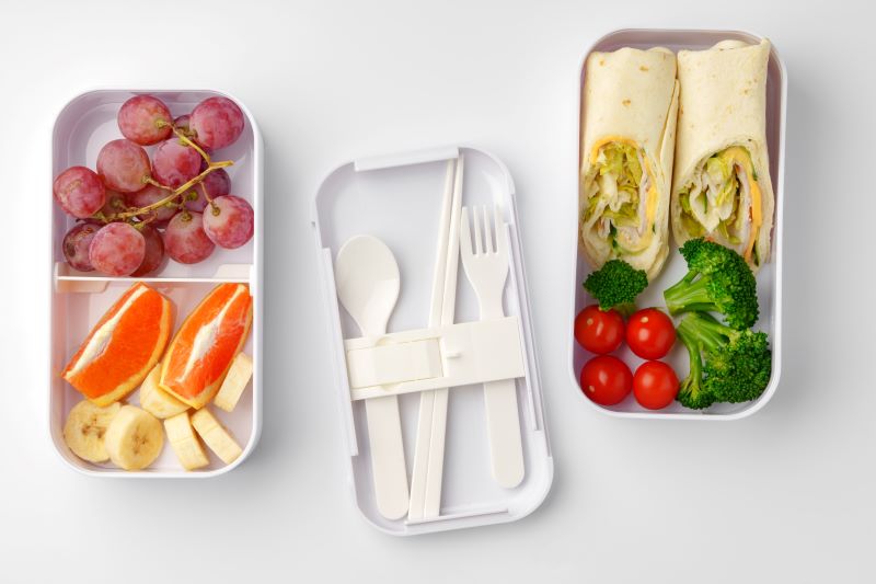 Two lunch boxes with fruit slices, vegetables, and wraps