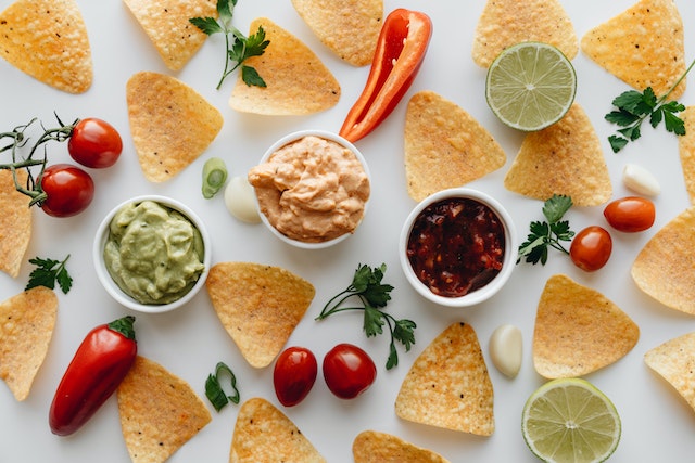 Nacho chips, dips, and spices, arranged on a white surface